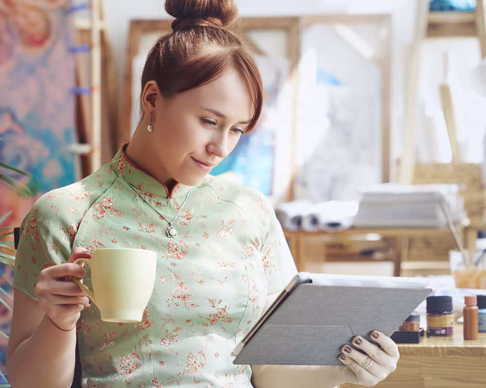 Junge Frau in einem Atelier hält eine Tasse und schaut auf ein Tablet, im Hintergrund Malutensilien und Staffeleien.