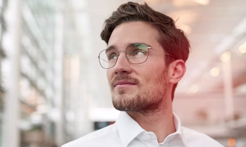 Mann mit Brille und Bart blickt nachdenklich zur Seite in einem hellen modernen Gebäude.